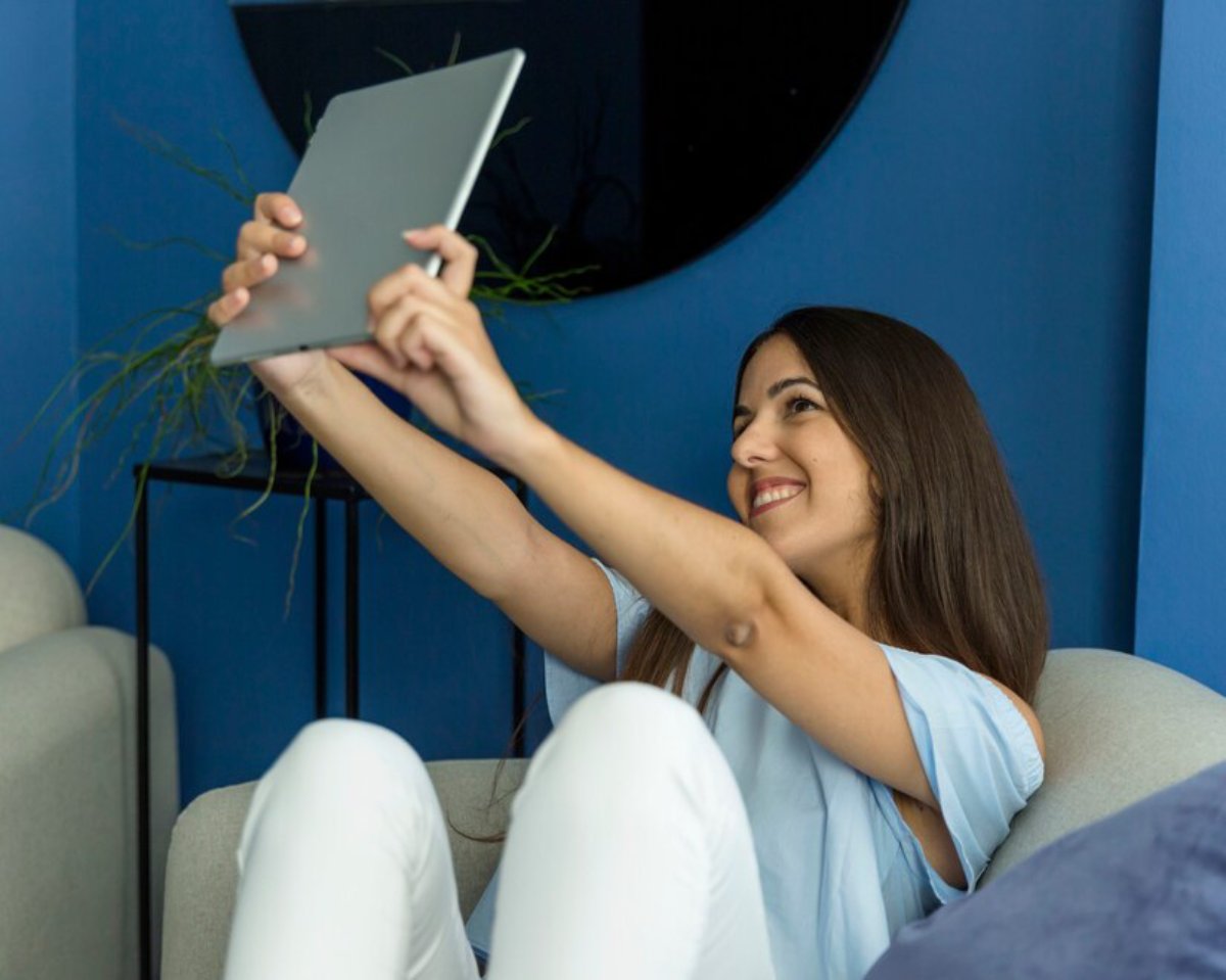 A person sitting on a couch, holding a tablet in front of them, in a modern room with blue walls and decorative plants.
