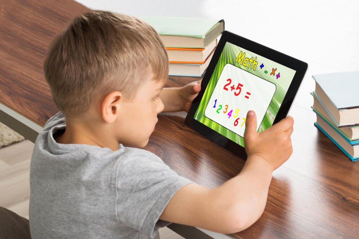 A child holds a tablet displaying a colorful math problem, surrounded by books on a wooden table.