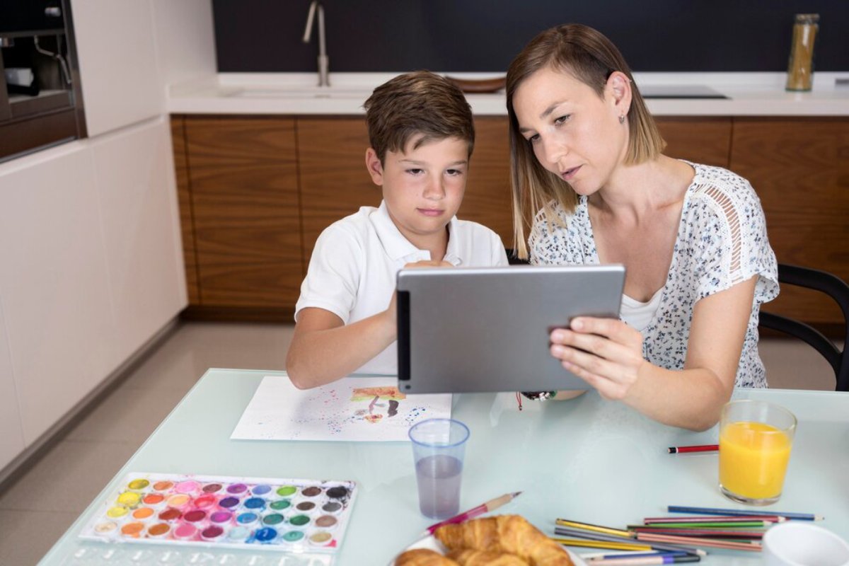 A woman and a child are sitting at a kitchen table, looking at a tablet. Art supplies and breakfast items are visible around them.