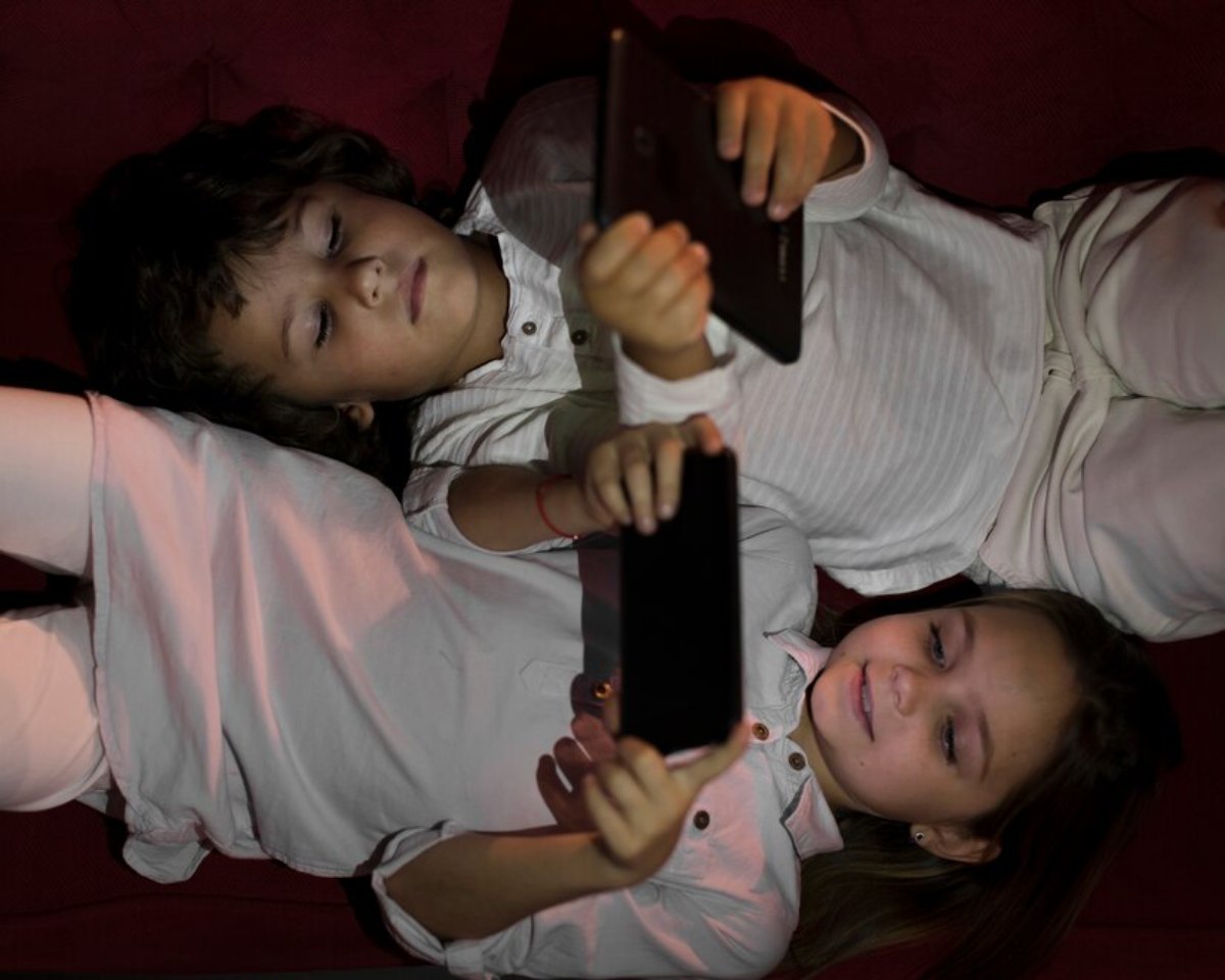 Three children lie on a couch, each engrossed in their electronic devices, dressed in white shirts and pants, creating a cozy scene.