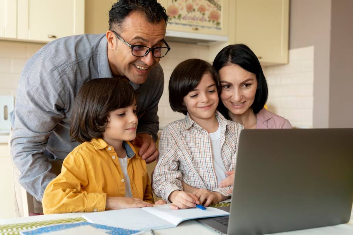 A family gathers around a laptop at a kitchen table, engaged in a shared learning experience with notes and a pen.