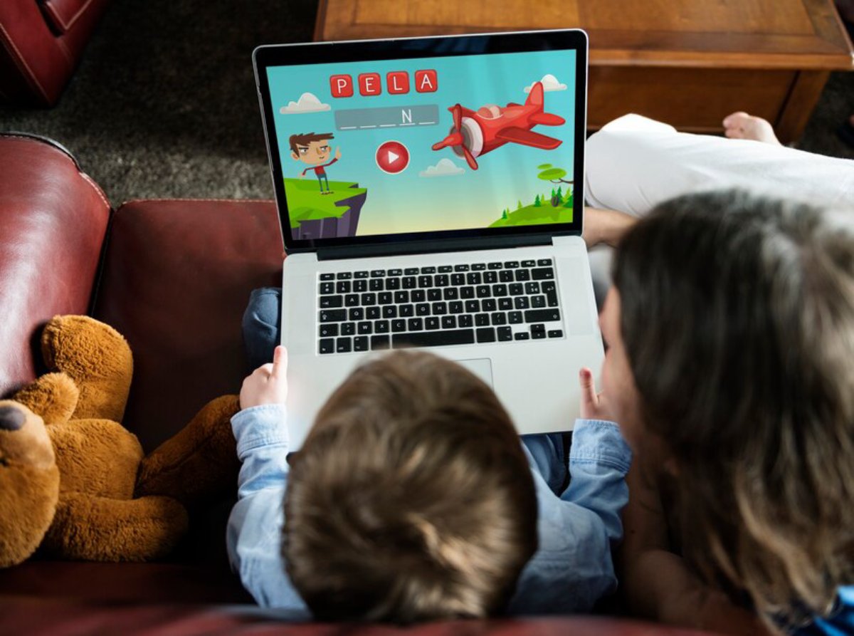 A child and a woman watch an animated educational video on a laptop, with a stuffed teddy bear nearby.