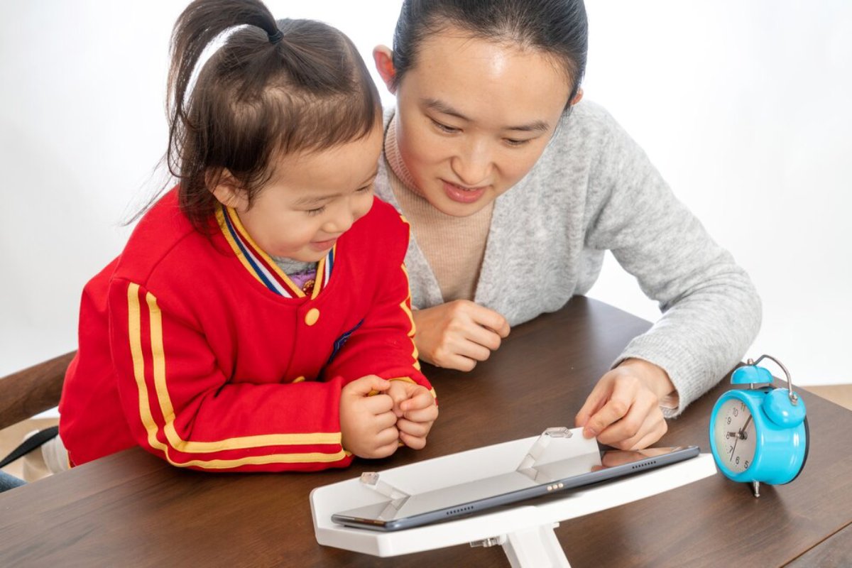 A child in a red jacket and an adult interact with a tablet on a wooden table, with a blue alarm clock nearby.