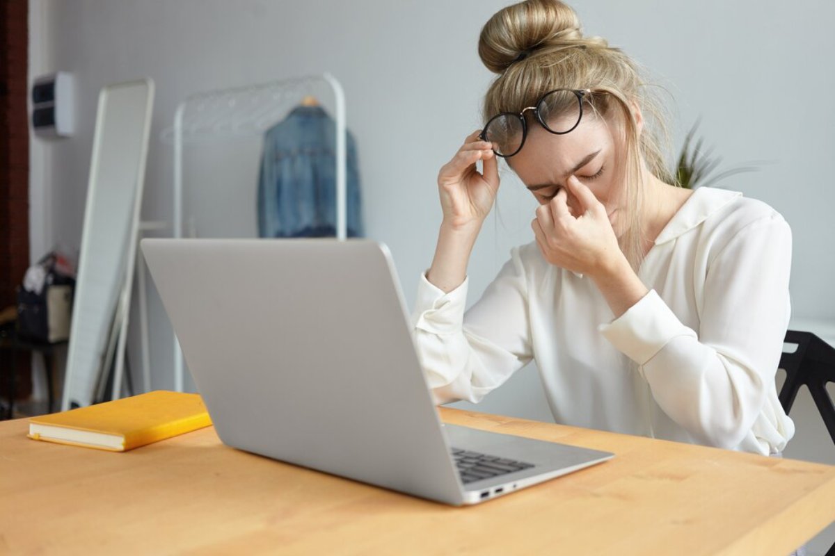 A woman in a white blouse sits at a wooden desk, focused on her laptop, adjusting her sunglasses. A yellow notebook is nearby.