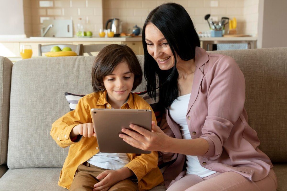 A woman and a child sit on a sofa, engaging with a tablet in a cozy kitchen setting filled with natural light.