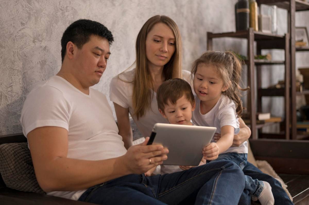 A family of four sits together on a couch, engaging with a tablet in a cozy living room setting.
