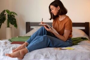 A woman sits on a bed, focused on writing in her journal, surrounded by soft pillows and warm lighting.