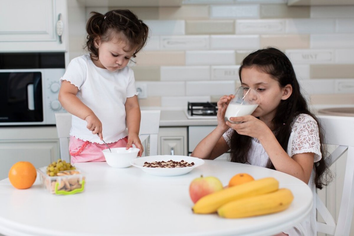 Two children are enjoying breakfast at a white table, featuring bowls of cereal, fruit, and a bowl of yogurt in a bright kitchen.