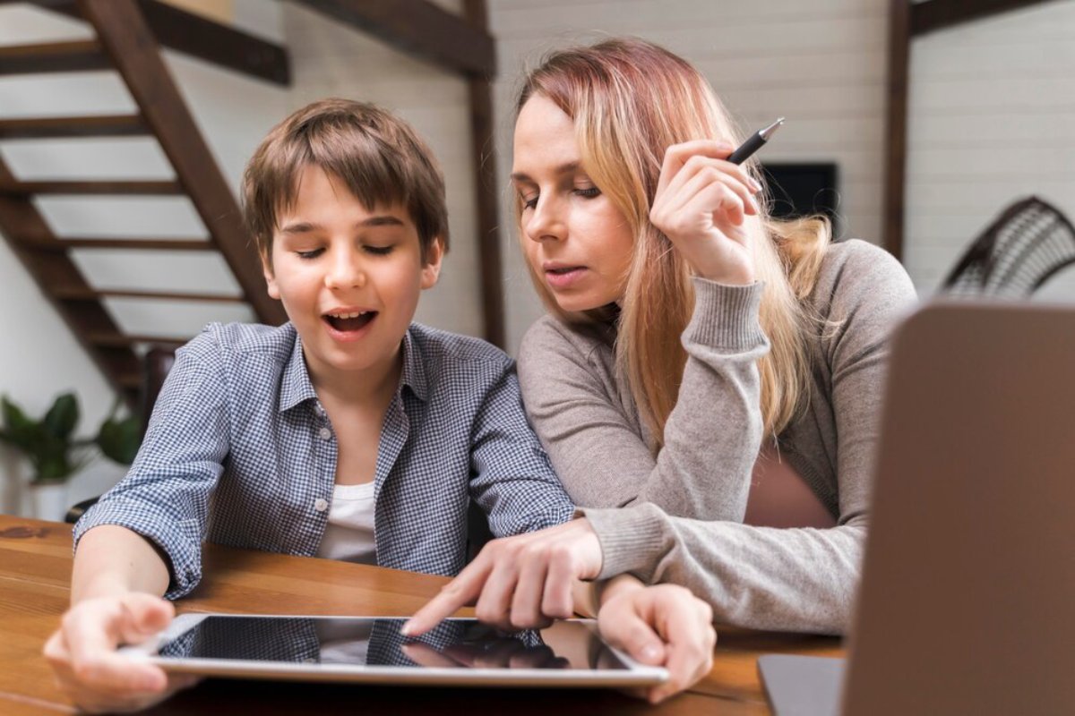 A child and a woman sit closely at a wooden table, engaged in learning and exploring a tablet together. A laptop is nearby.