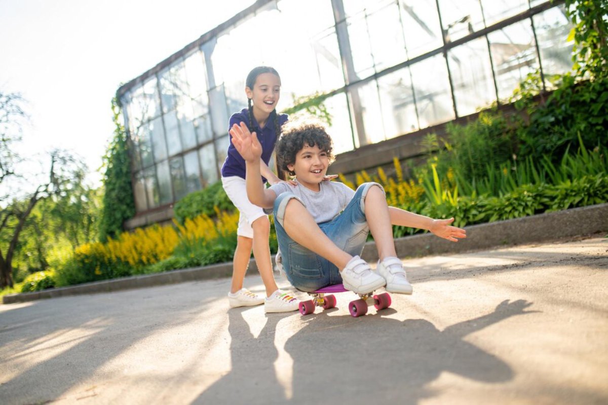 Two children play together outside, one skating while the other pushes, surrounded by greenery and bright sunlight.