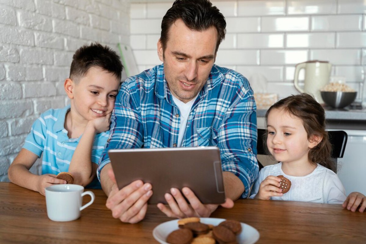 A man holds a tablet while sitting with two children at a wooden table, surrounded by cookies and a cup of coffee. Bright kitchen background.