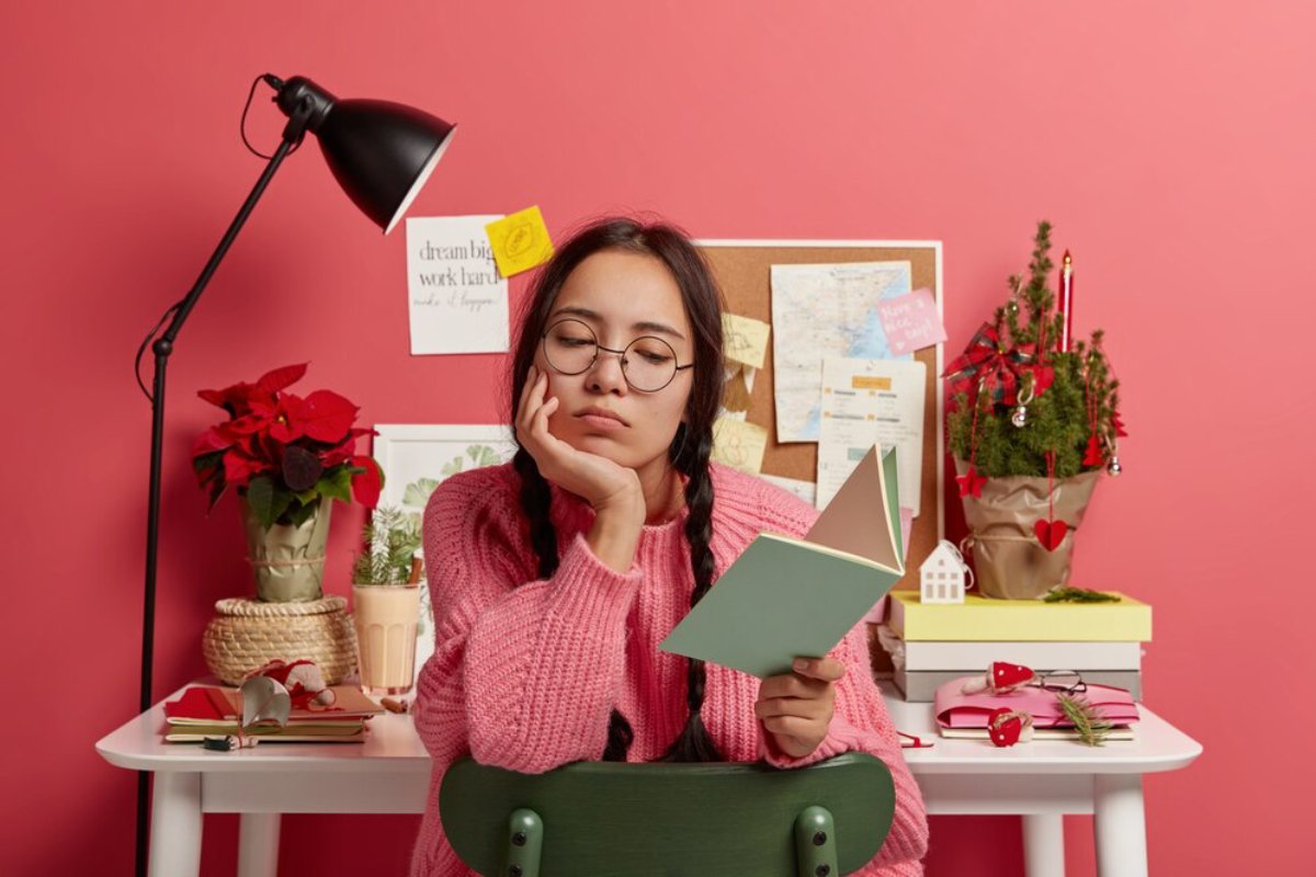 A young woman in a pink sweater sits at a green chair, holding an open book, surrounded by festive decor in a cozy workspace.