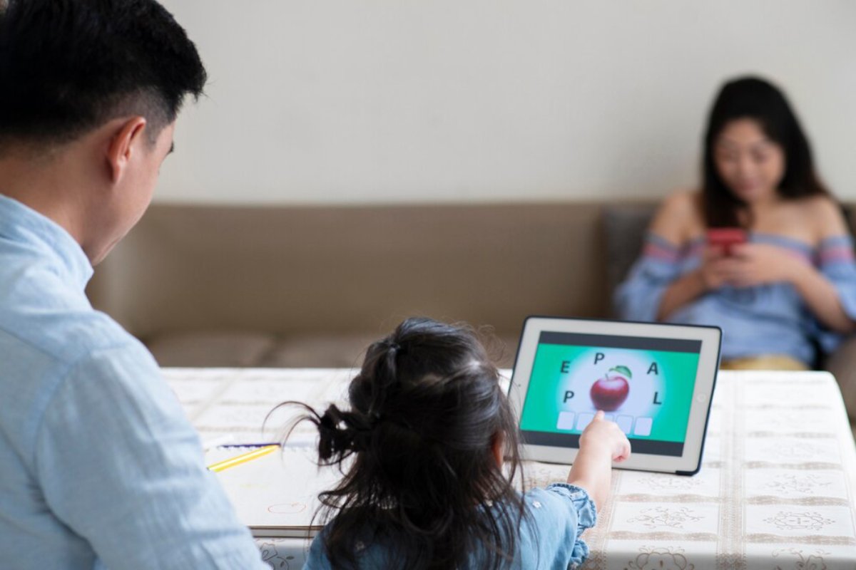 A young girl points at a tablet displaying an educational game, while an adult sits nearby, absorbed in a phone.