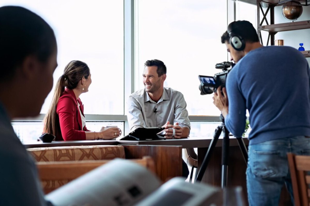 A man and woman having a discussion at a table during a filmed interview, with a cameraman and sound technician recording.