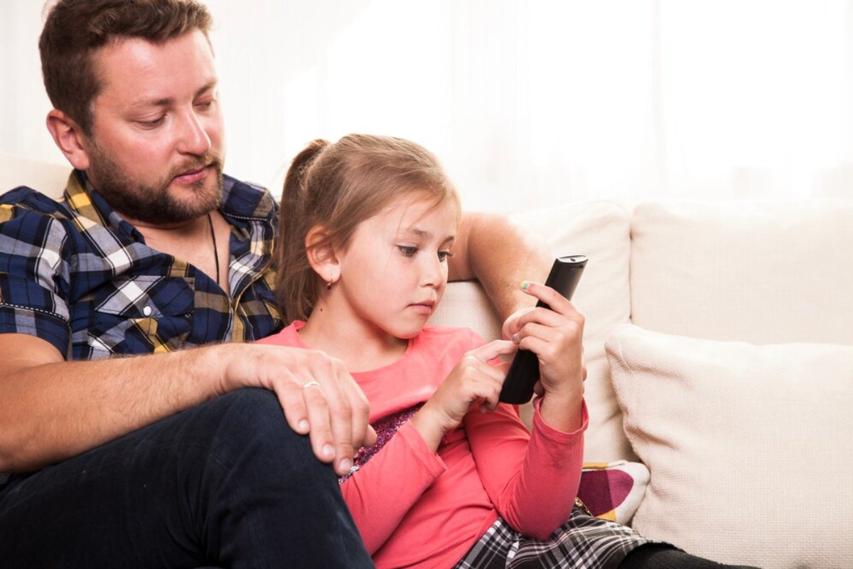 Father and daughter sitting on a sofa, looking at a smartphone together.