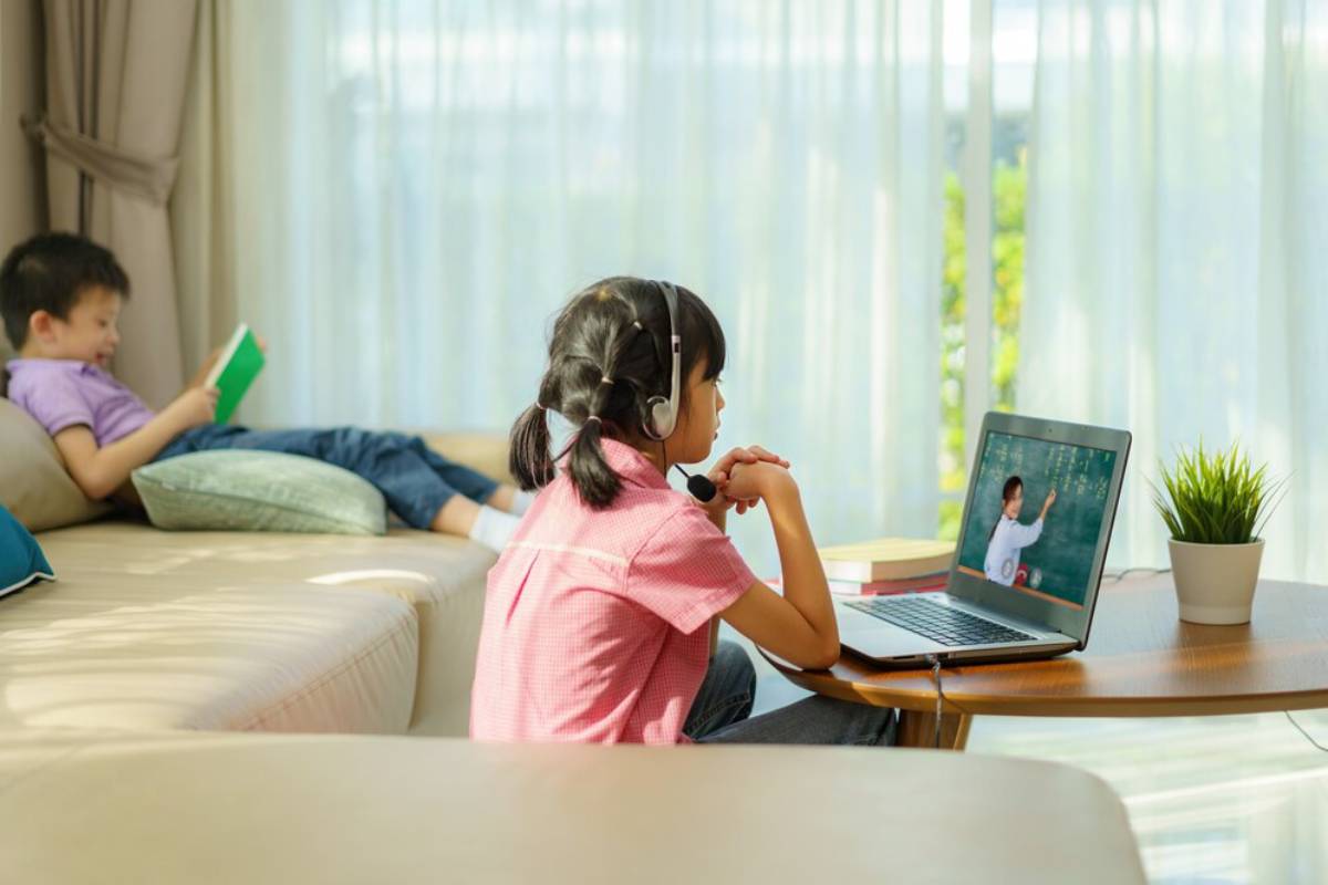 A young girl in headphones uses a laptop at a table while a boy reads on a couch in a sunny living room.