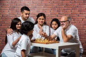 A multi-generational family plays chess together happily in front of a brick wall.