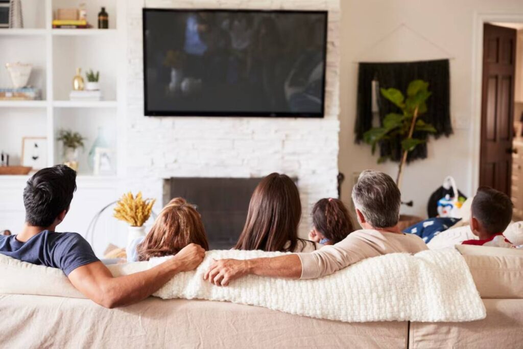 Family of five sitting on a couch, watching TV together in a cozy living room.