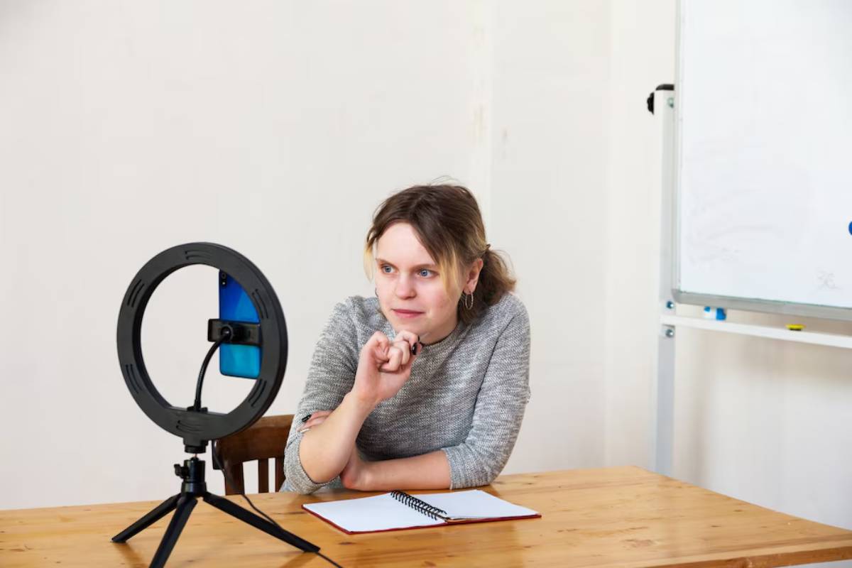 Woman sitting at a desk with a ring light and smartphone, appearing ready to record or stream.
