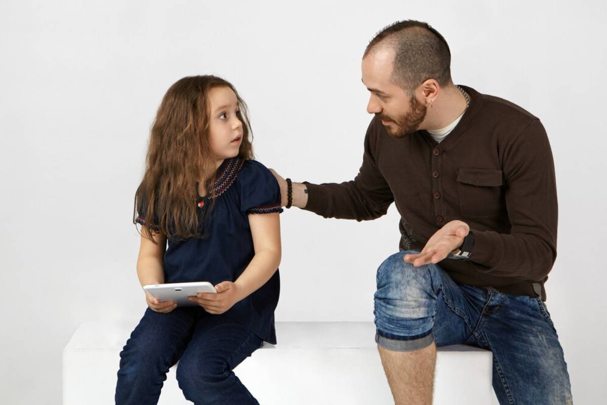 A man conversing with a young girl who is holding a tablet, both seated on a white surface.