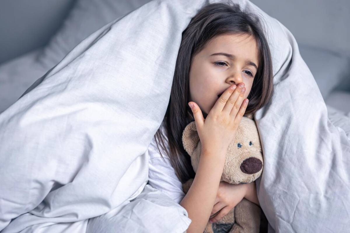 Young girl yawning in bed, holding a teddy bear, covered with a blanket.