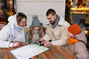 Family of four dressed in winter clothes looking at a camera and a map at an outdoor cafe table.
