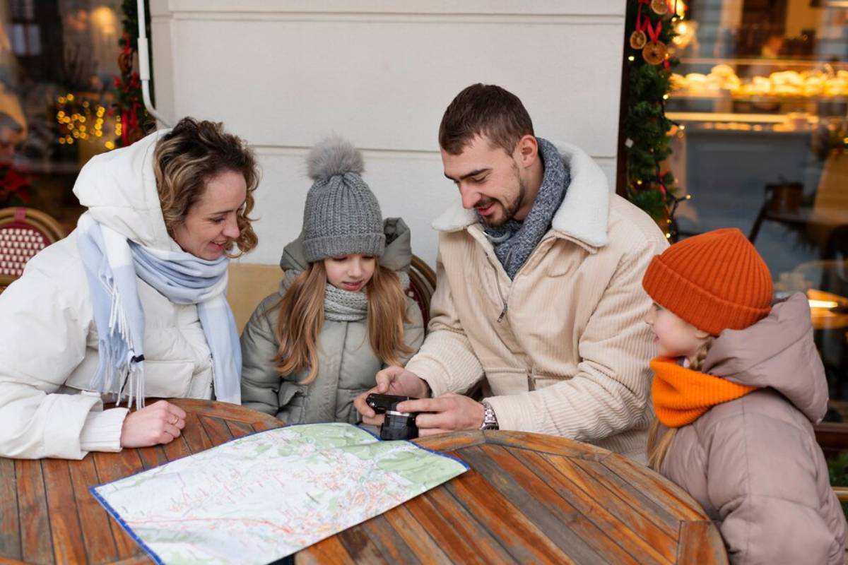 Family of four dressed in winter clothes looking at a camera and a map at an outdoor cafe table.