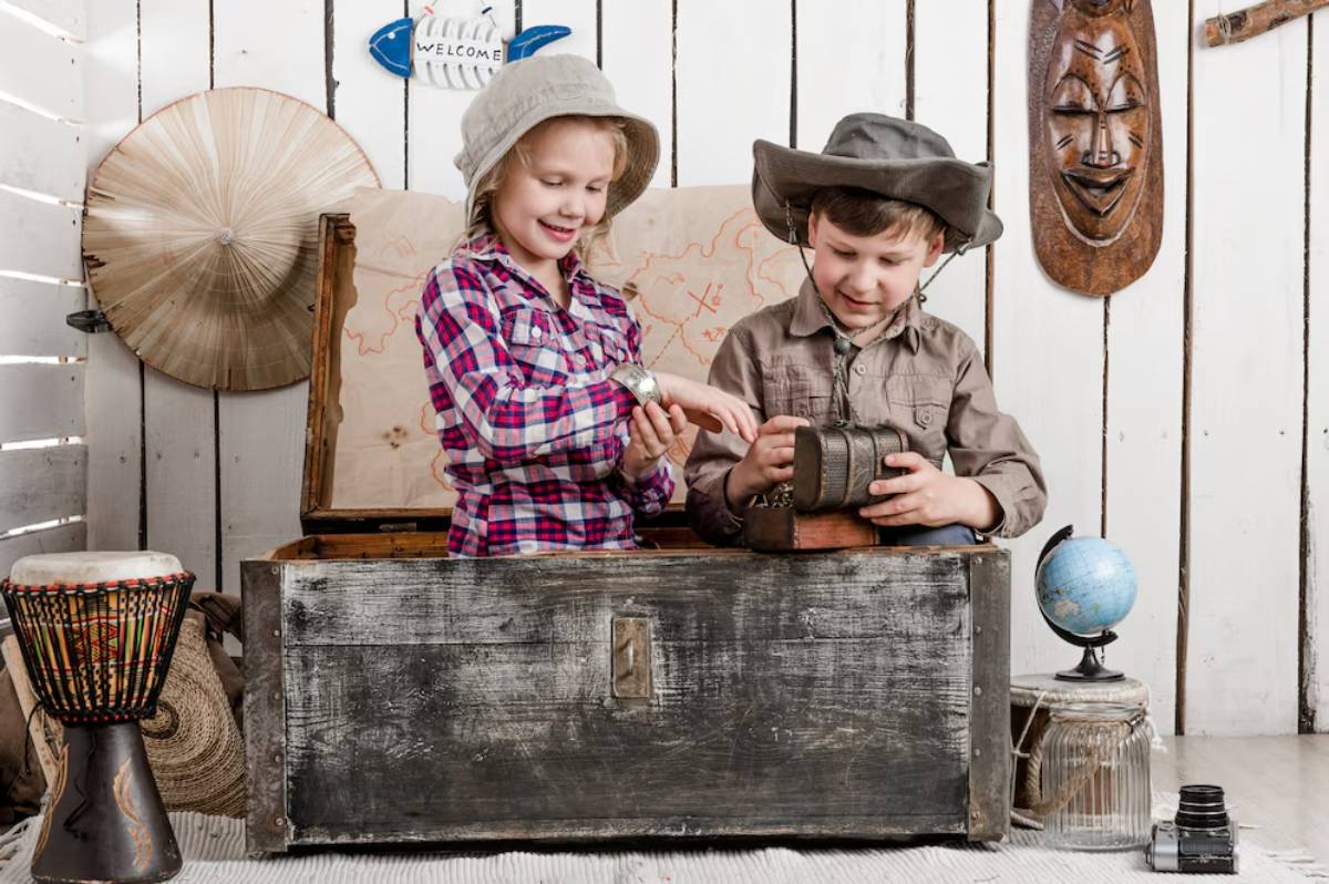 Two children dressed in explorer outfits examining old camera and artifacts near a wooden chest in a room decorated with travel-themed items.