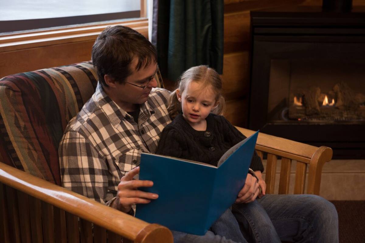 Father and young daughter reading a book together on a wooden bench by a fireplace.