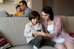A mother and son looking at a smartphone together on a couch while another child and man work on a laptop in the background.