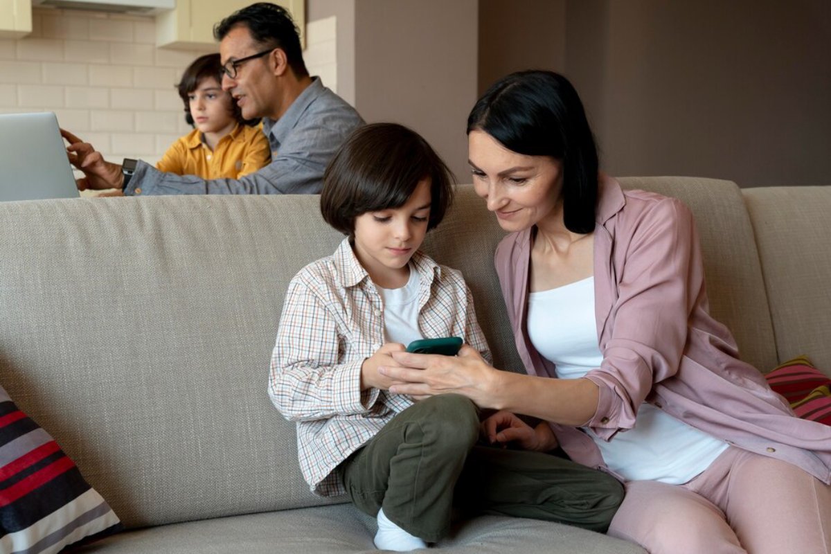 A mother and son looking at a smartphone together on a couch while another child and man work on a laptop in the background.
