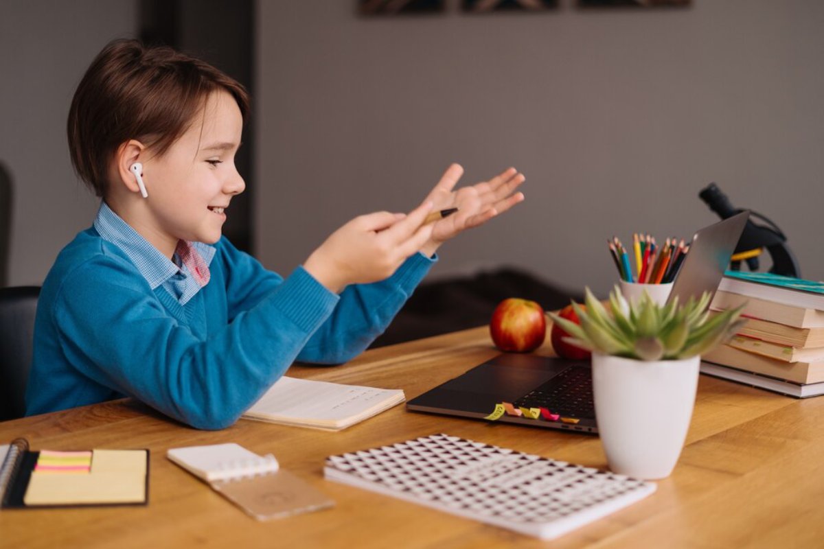 Young boy smiling while using a laptop and gesturing with his hands at a desk, surrounded by study materials.