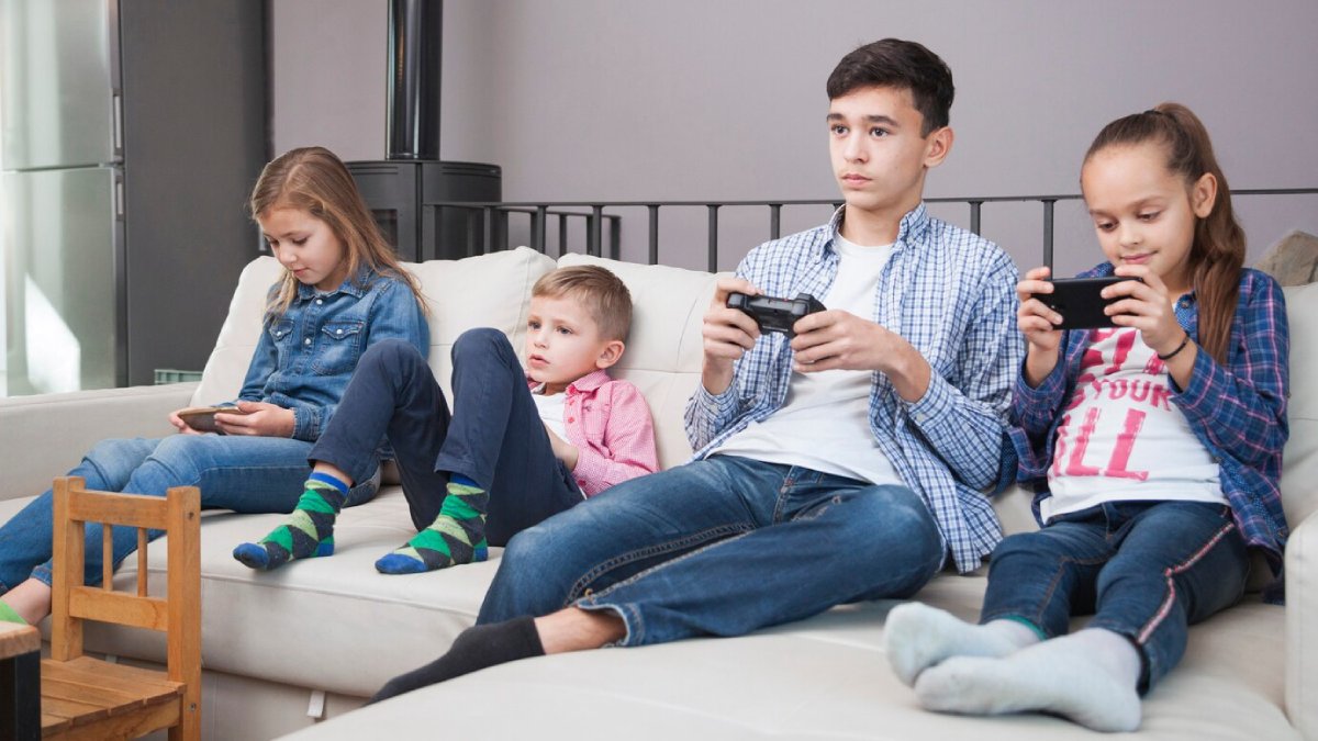 Four children sitting on a sofa, playing games on handheld devices in a modern living room.