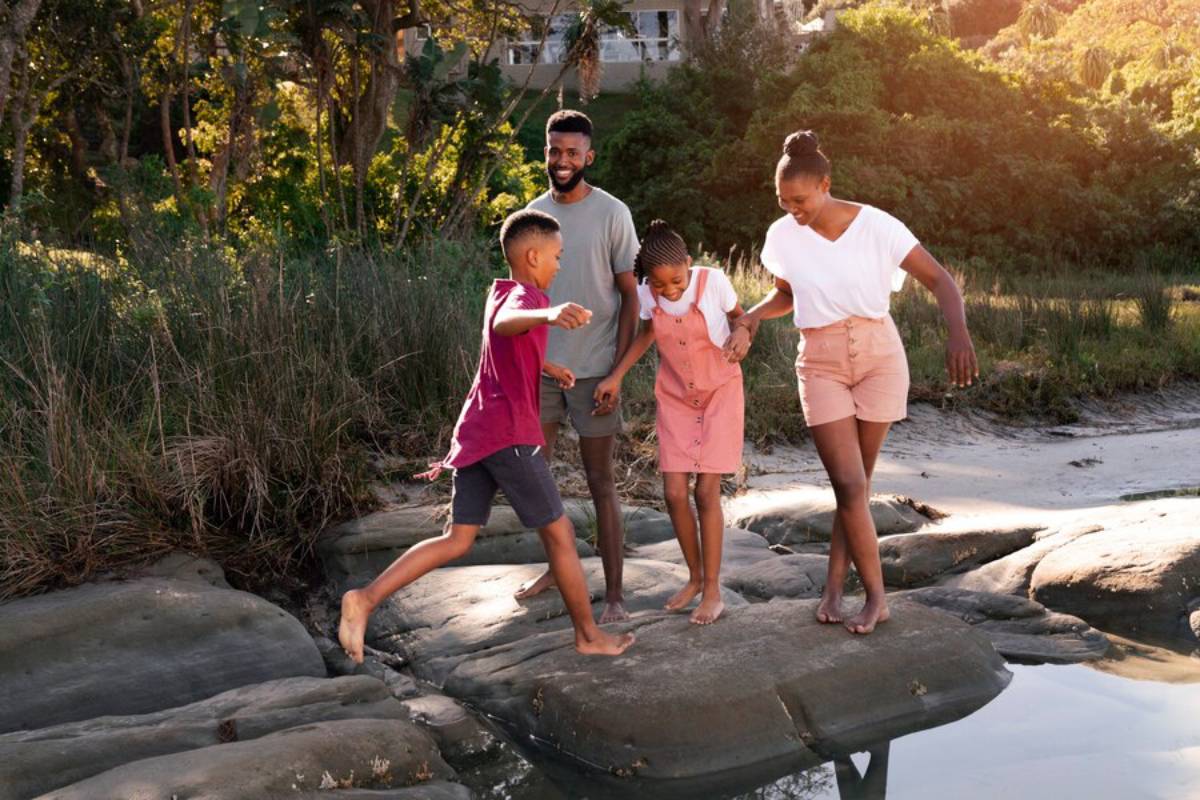 A family of four enjoying a playful walk on a rocky forest path near water.