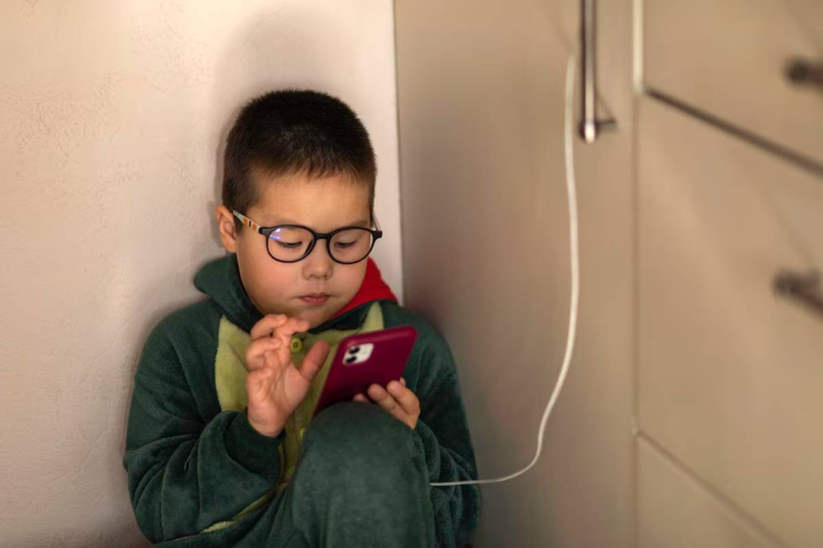 Young boy with glasses, focused on using a smartphone, sitting by a corner in a green hoodie.