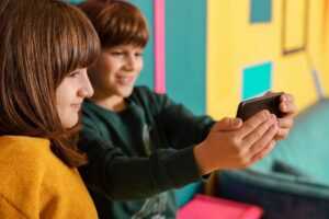 Two children smiling and looking at a smartphone together in a colorful room.