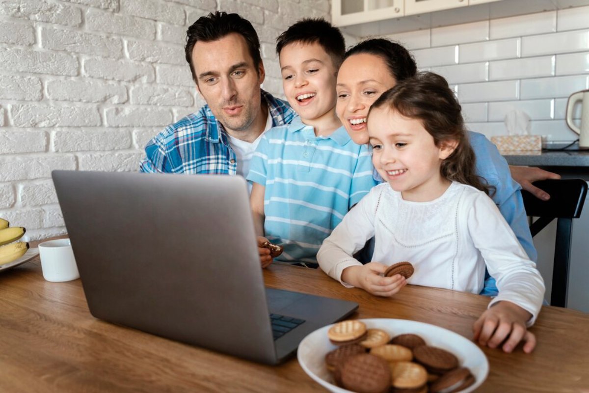 A family with two children excitedly watching a laptop in their kitchen, with a plate of cookies on the table.