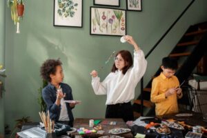 A woman and two kids craft with beads and paints in a cozy, art-filled room.