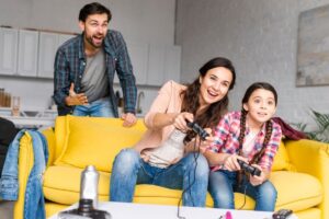 Family enjoying video games together; a woman and girl on a yellow sofa, and a man cheering in the background.