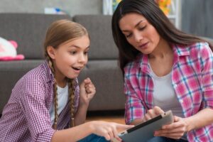 A woman and a girl sit on the floor, focusing intently on a tablet, sharing an engaging moment together in a cozy setting.