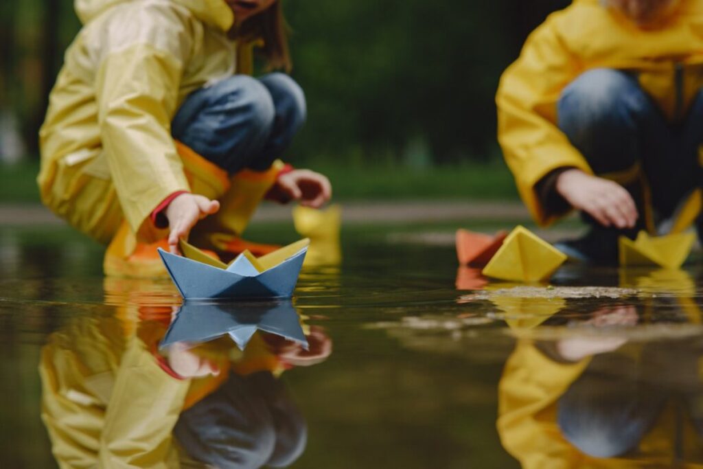 Children in yellow raincoats play with colorful paper boats in shallow water, creating ripples and reflections on the surface.
