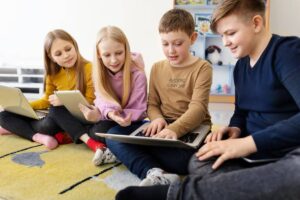 Four children sit on a rug, engaged in using tablets and a laptop, showcasing a focused and collaborative learning environment.