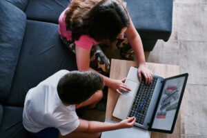Two children sit close together at a laptop, engaged in a project, with tools displayed on the screen and a notebook nearby.