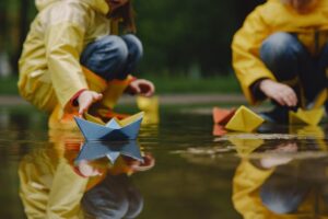 Children in yellow raincoats play with colorful paper boats in shallow water, creating ripples and reflections on the surface.
