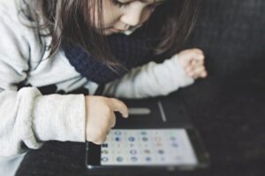 A person in a gray sweater uses a tablet with various app icons displayed, seated on a dark couch.