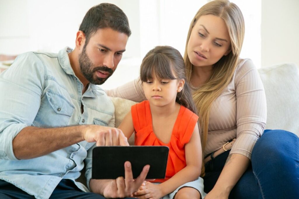A family sits together on a couch, engaged with a tablet, sharing a moment of connection and joy in a bright, cozy living room.