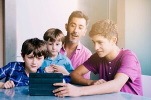A group of four children and a teenager sit around a table, focused on a tablet in front of them, immersed in digital content.