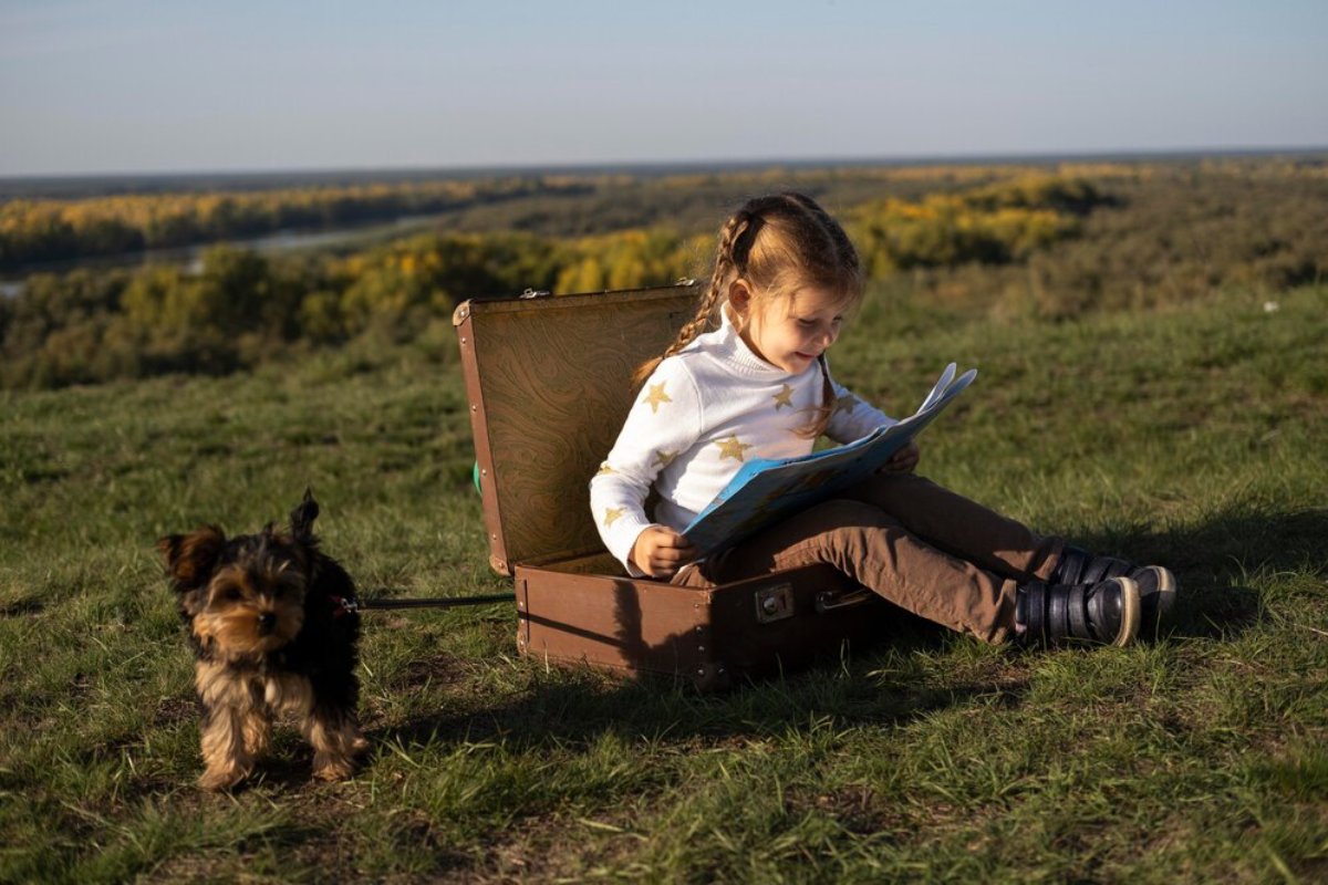 A child sitting on an open suitcase outdoors, reading a book, with a small dog standing nearby amidst a scenic landscape.
