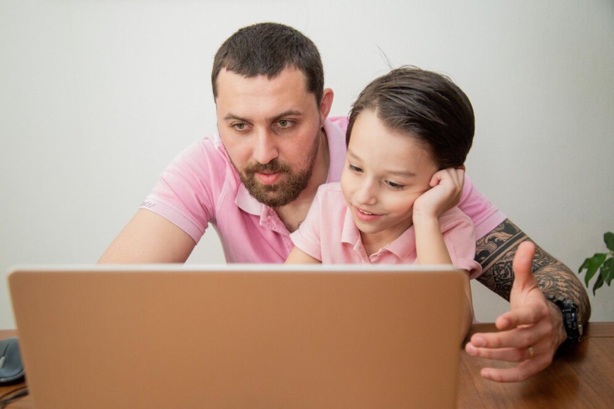 A man and a child in matching pink shirts sit closely together, focused on a laptop on a wooden table.