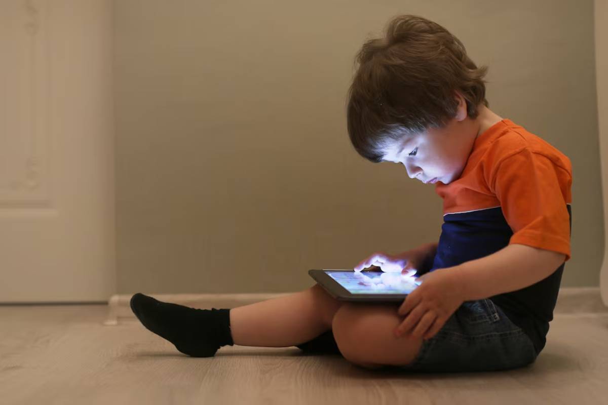 A young child sits on the floor, engrossed in a tablet, illuminated by its screen, wearing a colorful shirt and black socks.